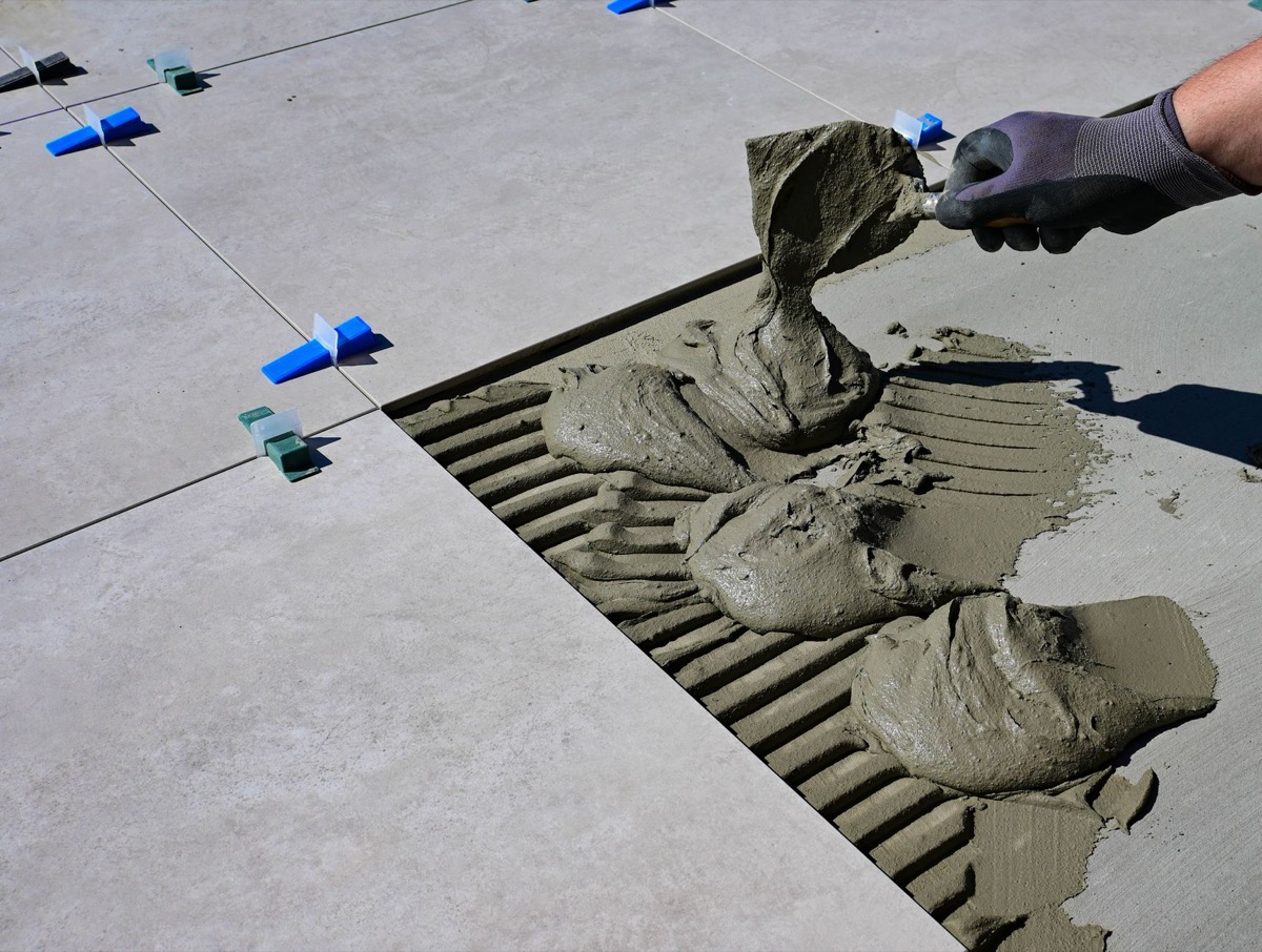 A person kneeling on a bathroom floor, spreading tile adhesive with a notched trowel and setting square ceramic tiles